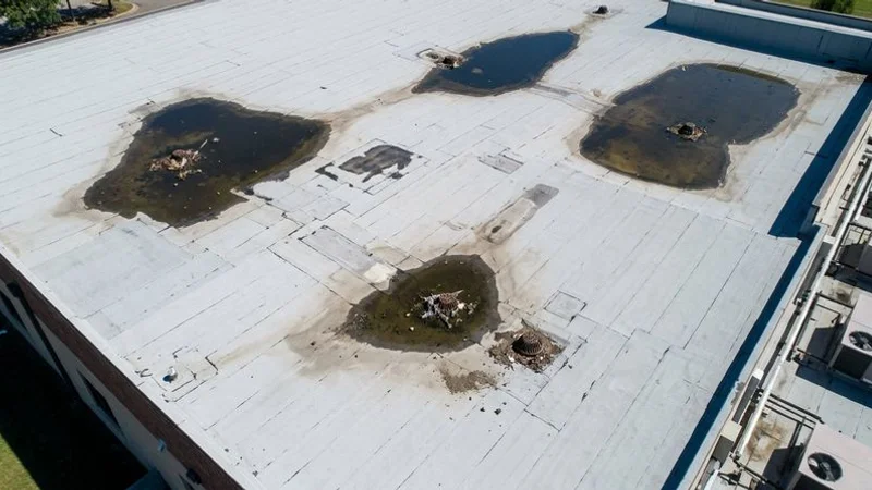 Aerial view of commercial flat roof showing multiple areas of ponding water with visible low spots and clogged drain locations needing repair